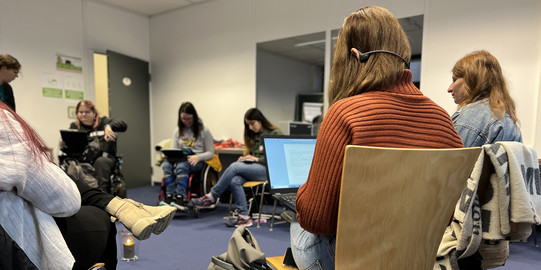 Studierende sitzen in einem Stuhlkreis zusammen, zwei Studierende sitzen im Rollstuhl, eine Studentin trägt einen Knochenkopfhörer, um den Screenreader ihres Computers zu hören und unter dem Stuhl einer anderen Studentin sitzt ein Blindenführhund.