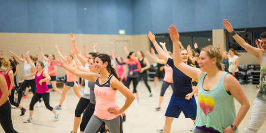 Teilnehmerinnen und Teilnehmer eines Fitness-Kurses in einer großen Sporthalle